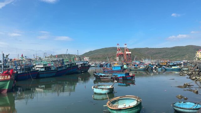 Traditional wooden fishing boats and round coracles moored in a calm harbor with modern industrial cranes in background.