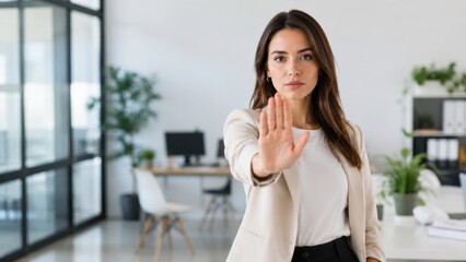Obraz premium Female business professional in a beige blazer holds up hand in a stop gesture, standing in a modern office with desks and plants in the background
