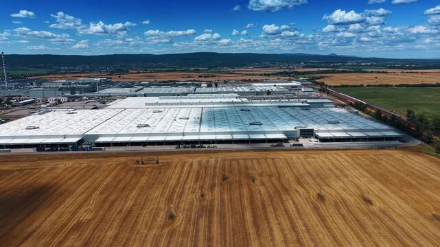 Large industrial complex beside harvested fields. White factory roofs extend across farmland with railway tracks, hills, and scattered clouds beyond.