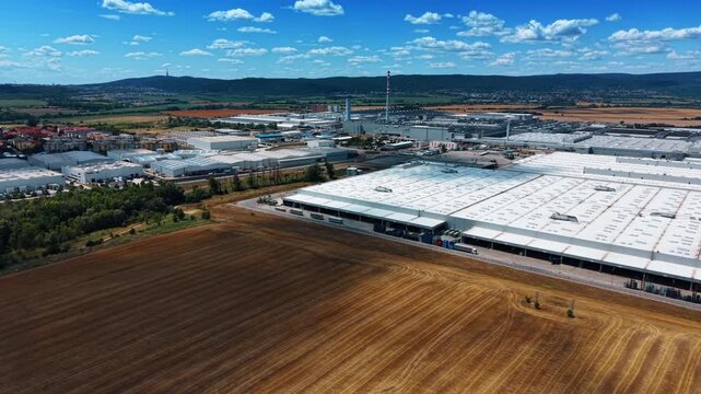 Factory district beside expansive harvested field. Large white halls and smaller workshops extend toward wooded hills under scattered summer clouds.