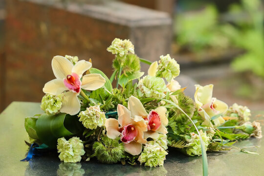 Memorial floral arrangement featuring greenish and yellowish flowers. Beautiful funeral flowers (carnations and orchids) on a memorial stone at the cemetery. An August day. Blurred background.