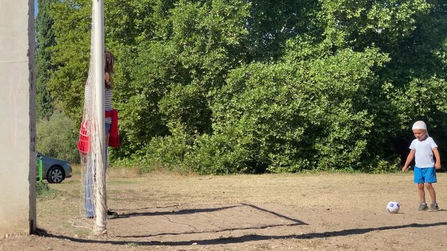 A young boy bends down to handle a soccer ball on a sunny field as a woman stands watching near the goal. They enjoy a peaceful, joyful afternoon together surrounded by green trees.