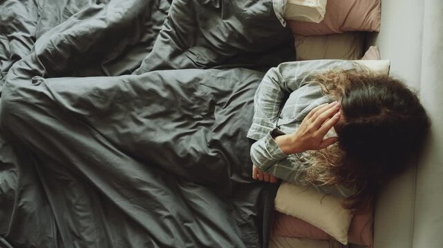 sad face lonely unhappy young woman sitting alone in bed, waking up with anxiety depression poor mental health, headache. top adobe view upset stressed tired girl cry sleep problems insomnia migraine.