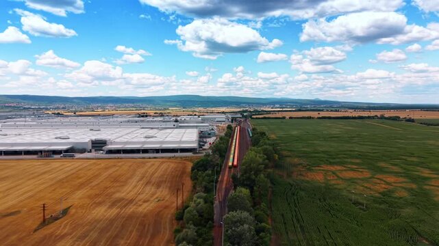 Railway line separating industrial and agricultural land. A train on multiple tracks runs between a large white warehouse building and a green cultivated field.