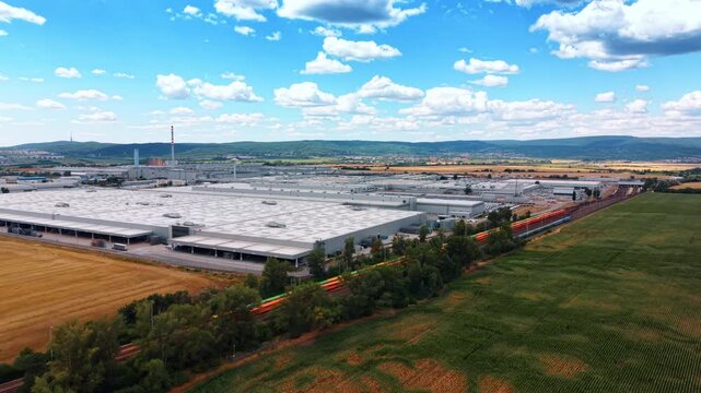 Aerial view of industrial complex bordered by farmland. Factory buildings extend beside a tree lined railway, bordered by golden wheat and green cornfields under cloudy skies.
