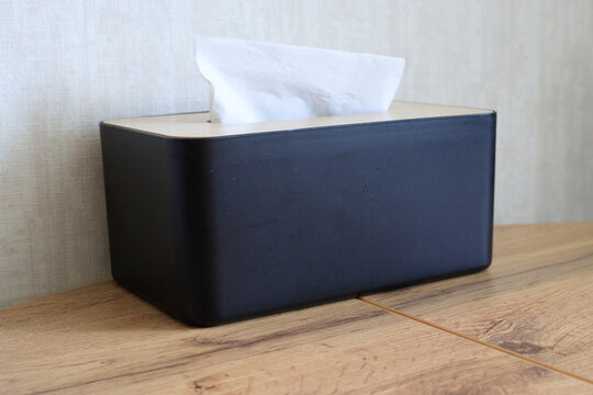 Black, minimalist paper napkin container sits on a table. White paper napkin protrudes from the black tissue box. Modern hygiene concept