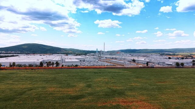 Aerial establishing shot of vehicle factory and parked cars. Rows of new vehicles line a factory beside a railway track, framed by rolling hills and thick white clouds.