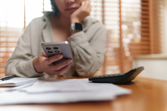 Asian Woman checks financial information on a smartphone while reviewing bills and a calculator, highlighting budgeting, expense management, and personal financial planning at home.