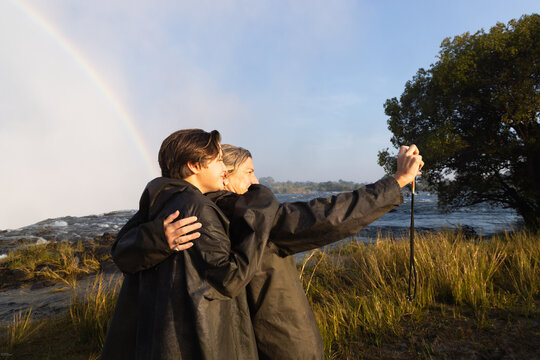 mother and her son taking a selfie , edge of Zambezi River as it drops into Victoria Falls, Zambia, Africa, rainbow in background