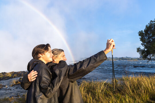 mother and her son taking a selfie , edge of Zambezi River as it drops into Victoria Falls, Zambia, Africa, rainbow in background