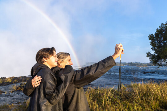 mother and her son taking a selfie , edge of Zambezi River as it drops into Victoria Falls, Zambia, Africa, rainbow in background