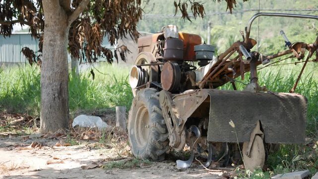 A small old tractor sits in a rural field beside a tree, with grass and open land in the background. Farm equipment.