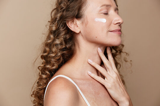 Authentic beauty of a middle aged woman with natural skin, applying moisturizer on her cheek. Close up portrait with warm studio light and soft beige background