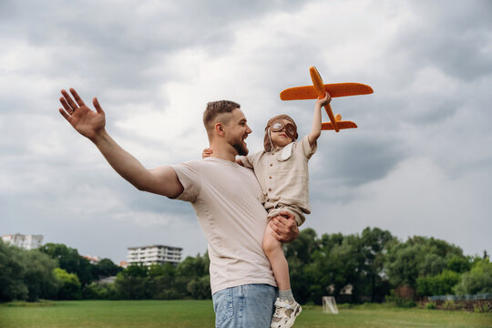 Father holding child with toy airplane in a green park, both smiling and enjoying a playful moment outdoors under a cloudy sky
