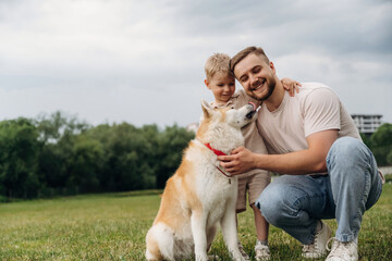 Obraz premium Caucasian father and son kneeling on grass petting a friendly Akita dog in a green park with trees and a cloudy sky in the background