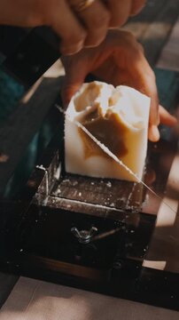 close up shows hands guiding handmade soap through wire cutter, revealing creamy swirls and warm light