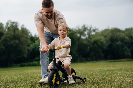 Father assists young boy riding a tricycle on grassy field, surrounded by trees and open space, showcasing a playful outdoor moment in a natural setting