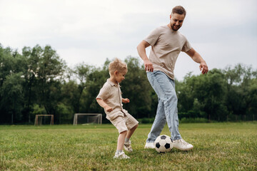 Obraz premium Father and son playing soccer on grassy field, child in beige outfit and father in casual wear, soccer ball at their feet, goalposts visible in the background