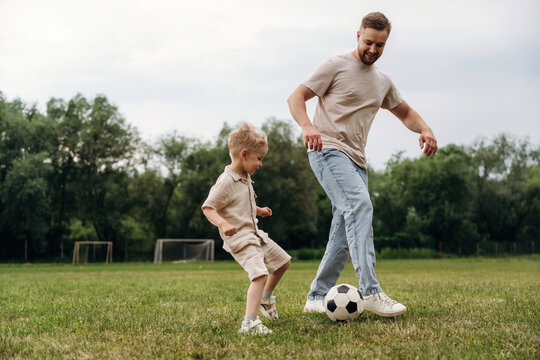 Father and son playing soccer on grassy field, child in beige outfit and father in casual wear, soccer ball at their feet, goalposts visible in the background