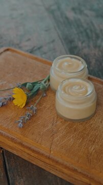 close view shows two cream jars on scratched wooden board with marigold and lavender petals around