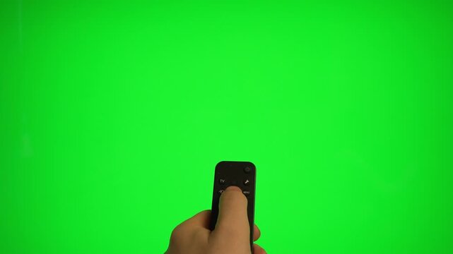Hand holding remote control pointing at green screen TV mockup.