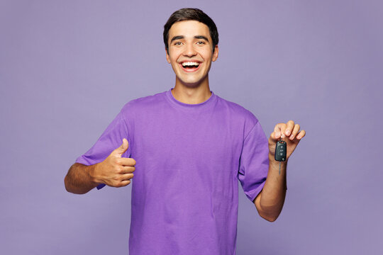 Young happy smiling fun cool man wear violet t-shirt casual clothes hold car key fob keyless system show thumb up isolated on plain pastel light purple background studio portrait. Lifestyle concept