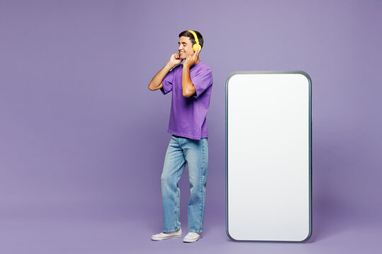Full body young man wear violet t-shirt casual clothes big huge blank screen mobile cell phone smartphone with workspace area listen to music in headphones isolated on plain light purple background.