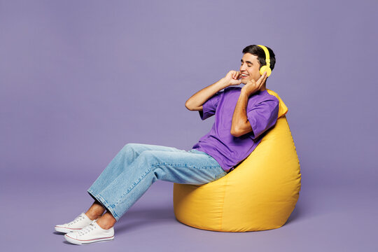Full body young man he wear violet t-shirt casual clothes sit in bag chair listen to music in headphones close eyes isolated on plain pastel light purple background studio portrait. Lifestyle concept.