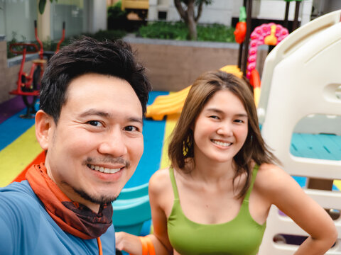 Happy Asian couple smiling for a selfie at an outdoor playground, enjoying leisure time together.