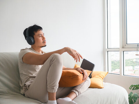 Thoughtful Asian Man Wearing Headphones Relaxing on Sofa at Home, Looking Out Window