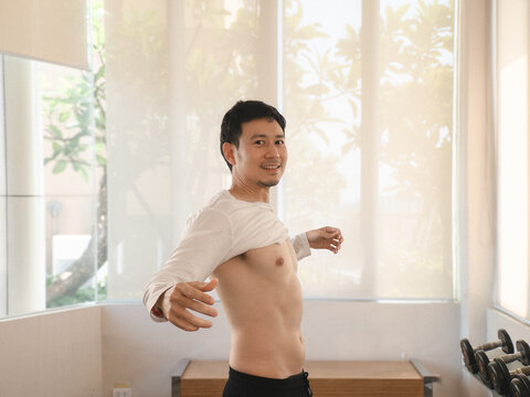 Fit Asian man smiling, confidently taking off his shirt in a bright gym.