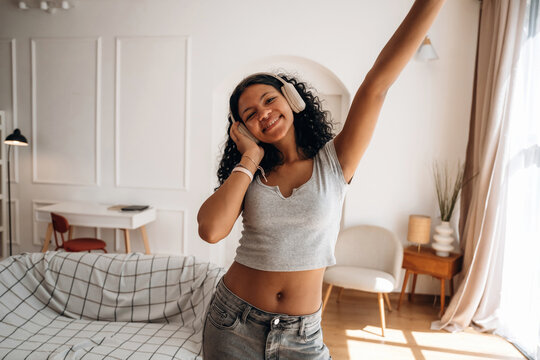 Young woman with curly hair wearing headphones dances joyfully in a bright living room with a cozy sofa and modern furniture, sunlight streaming through the window