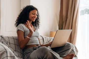Young woman with curly hair sitting on a couch using a laptop, waving and smiling, with natural light streaming through a window in a cozy living room setting © standret