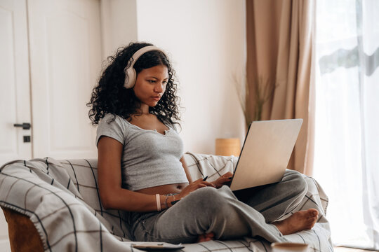 Young African woman with curly hair wearing headphones sits on a couch using a laptop in a bright, modern living room with natural light streaming through the window