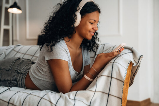 Young woman with curly hair wearing headphones uses smartphone while lying on a cozy couch with a plaid blanket in a bright indoor living space