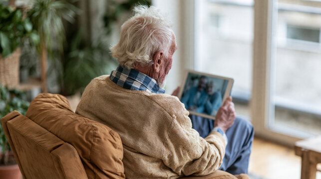 Rear view of a senior man holding a tablet during a video call while relaxing in an armchair beside a bright window at home.