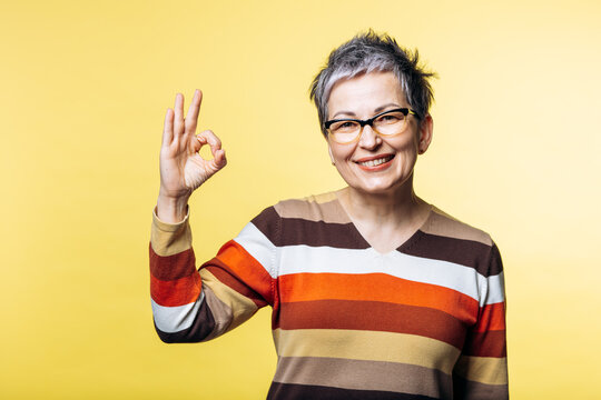 Happy senior woman confidently showing an okay hand gesture, expressing approval and positivity against a yellow background