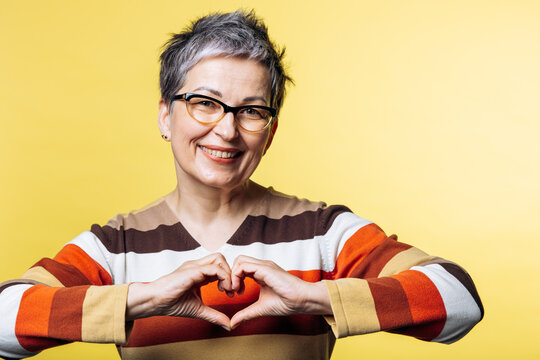 Cheerful senior woman with grey hair and glasses smiling, expressing love and affection with a heart hand gesture