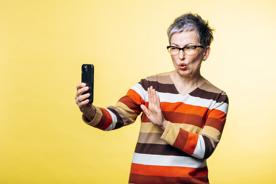 Senior woman posing with a funny face, holding a smartphone and taking a selfie against a yellow background