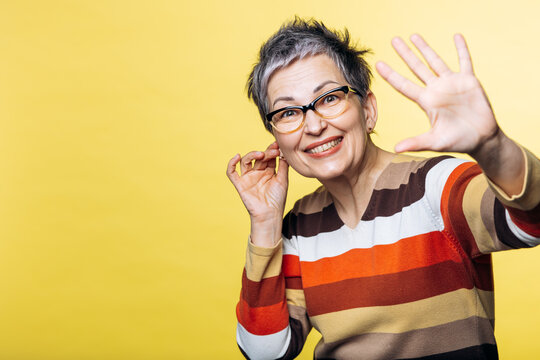 Happy mature woman with short grey hair and glasses covering face with hand, wearing striped sweater on yellow background