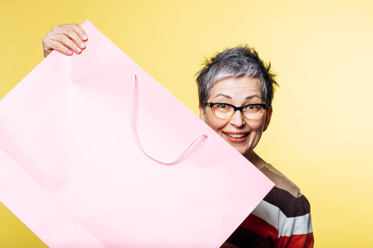 Happy senior woman holding a large pink paper shopping bag, peeking and smiling at the camera on yellow background with copy space
