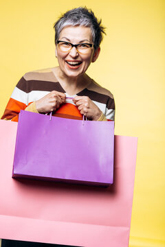 Cheerful mature woman with short grey hair and eyeglasses enjoying shopping, holding colorful bags on a yellow background