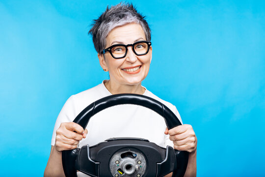 Happy mature woman smiling, holding a car steering wheel against a blue background. Representing driving lessons and independence