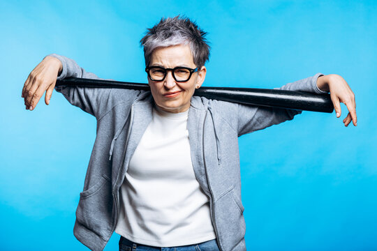 Confident senior woman in glasses and hoodie holding a baseball bat on shoulders, standing against a blue background