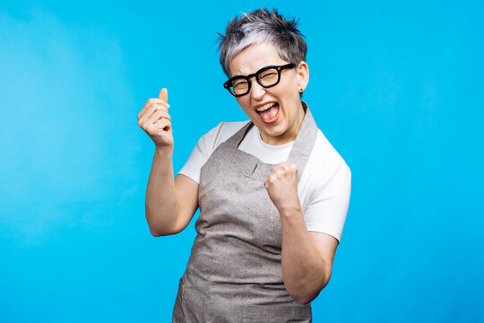 Woman in an apron exulting with fists clenched and mouth open, celebrating achievement and victory against a blue background