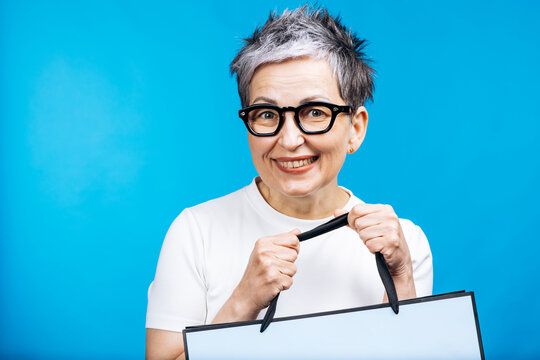 Cheerful older woman with glasses holding a shopping bag, smiling towards camera against a bright blue background