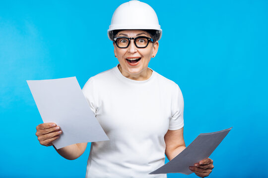 Senior woman engineer wearing a hard hat and glasses, looking surprised and pleased while holding and reviewing documents
