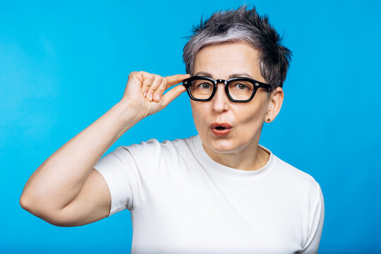 Mature woman making a surprised facial expression, adjusting her glasses with one hand against a blue background