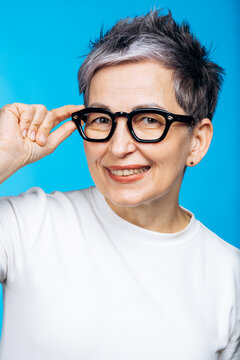 Mature woman with gray hair wearing a white sweater, adjusting her black eyeglasses and smiling at the camera on a blue background