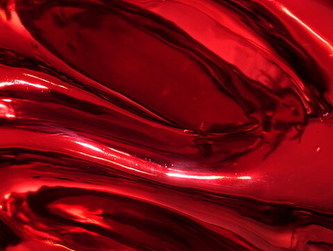 Close-up of a glossy, reflective red metallic surface with smooth fluid waves, bright highlights, and deep shadows creating an abstract, dynamic texture.